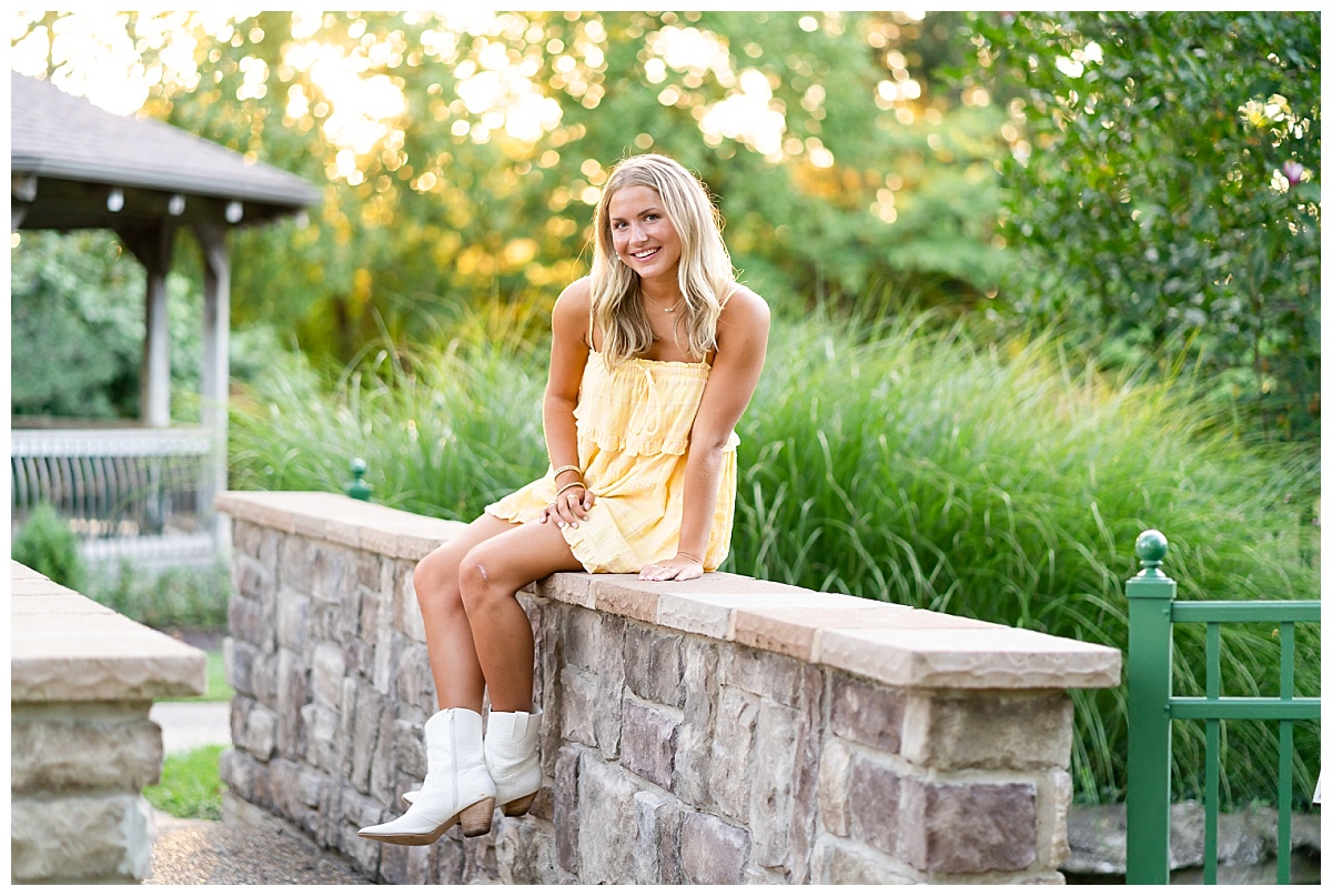 senior girl wearing yellow romper and white cowgirl boots poses on a bridge for her senior session with Hannah Carr Photography at Shelter Gardens in Columbia, MO
