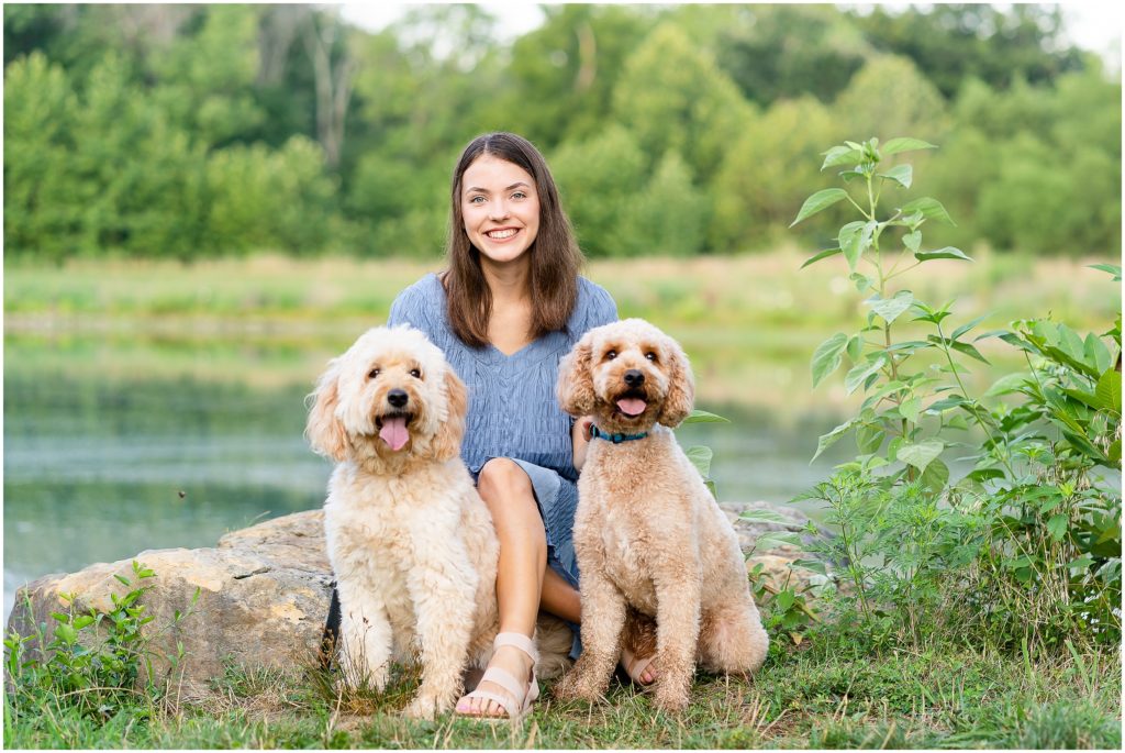 Senior girl poses with her dogs on a rock by a pond at Nifong Park in Columbia, MO for her senior portrait session with West Plains, MO photographer, Hannah Carr Photography