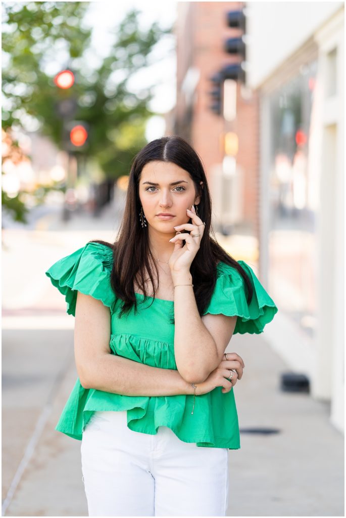 Senior girl poses in a green flowy top and white pants on a sidewalk downtown Columbia Missouri for her senior session with West Plains Photographer, Hannah Carr Photography.