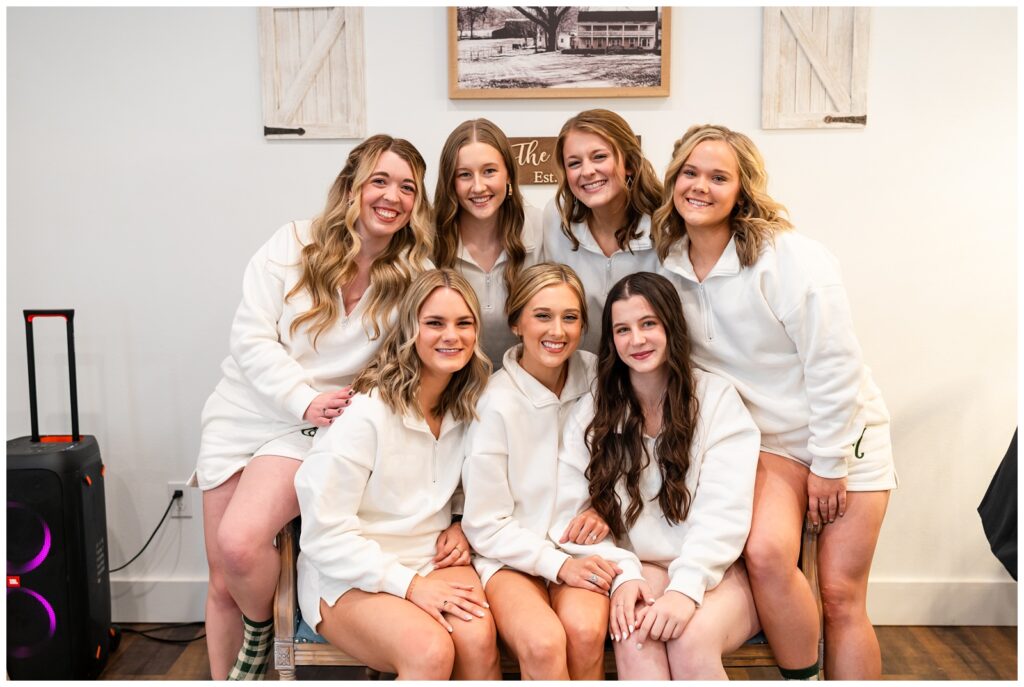 A photo a bride and her bridesmaids in their getting ready attire posing on a bench in the bridal suite of The Elms Barn in Flippin, AR taken by Arkansas wedding photographer - Hannah Carr Photography.