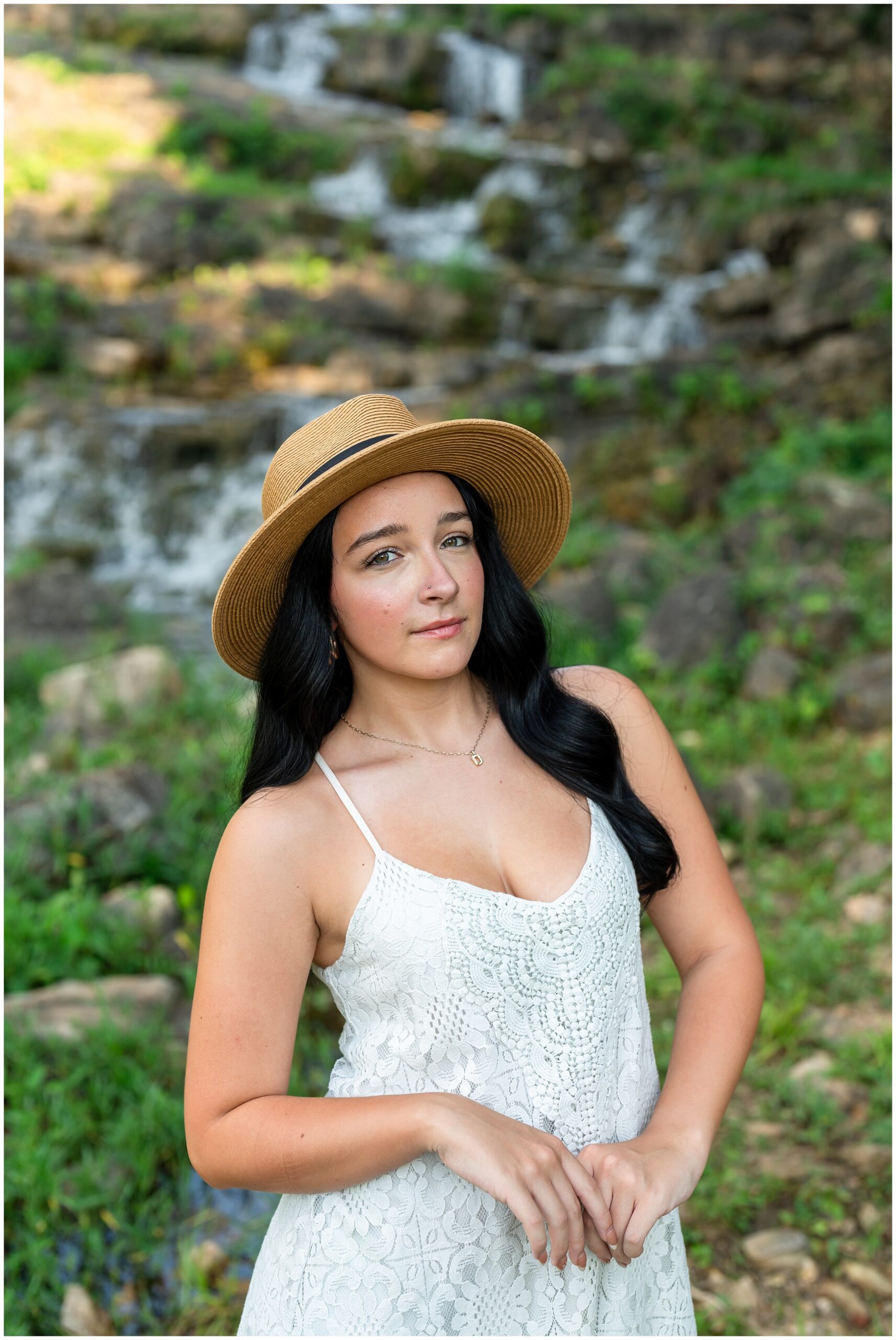 Highland High School senior, Dacy-Lynn, poses near a water fall wearing a white dress and hat in beautiful Cherokee Village, AR for her senior session with North Arkansas Senior Photographer, Hannah Carr Photography.